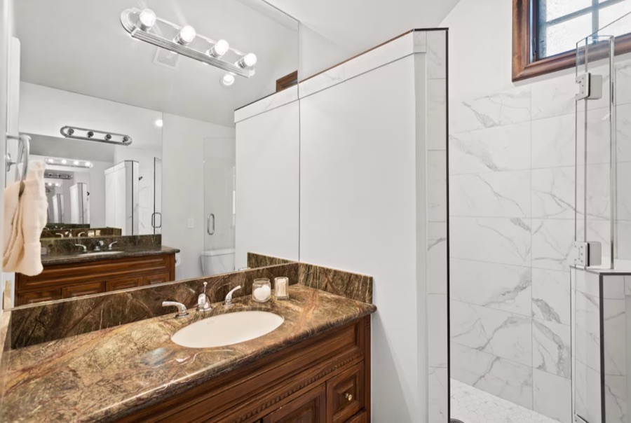 Bathroom with a granite-topped wood vanity, large mirror and a glass-enclosed marble-tiled shower.